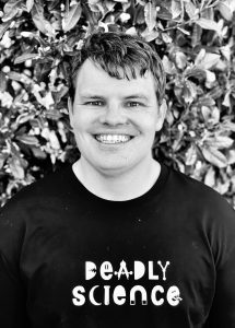 A greyscale headshot of Corey Tutt in front of a hedge, wearing a shirt with the text: Deadly Science.