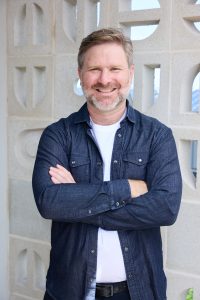 Portrait of Ed Coper standing with arms folded in front of a wall of decorative concrete blocks.