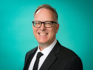 Studio portrait of Garth Nix standing at an angle and smiling, against a turquoise backdrop.