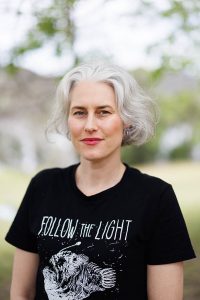 Outdoor headshot of Eleri Harris wearing a black T-shirt printed with 'follow the light'.
