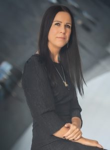 Headshot of Irma Gold wearing a black dress before an abstract grey background.