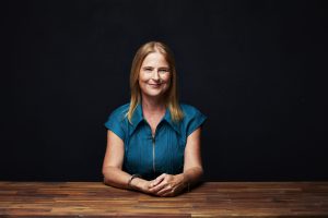 Studio portrait of Jaclyn Moriarty seated with hands clasped against a dark background.