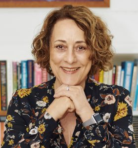 Headshot of Jane Messer smiling with clasped hands under chin, in front of shelves of books.
