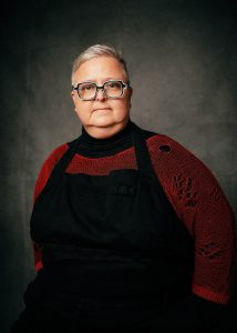 Moody studio portrait of Kris Kneen looking into the camera with their hands in their pockets. They are in front of a gray background.