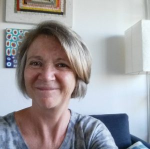 A selfie of Linda Wells sitting on a couch with paintings behind her. She is smiling.