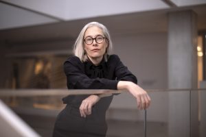 A portrait of Sophie Gee in front of a grey pillar, with her arms resting on a glass barrier.