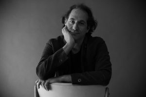A dramatic greyscale studio portrait of Steve Toltz sitting backwards on a chair.