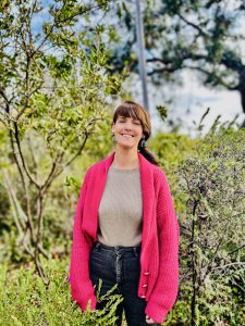 Outdoor portrait of Jennifer Pinkerton, smiling, in a hot pink cardigan in a bush setting.
