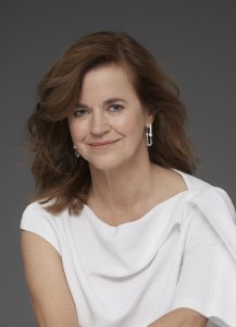 A studio portrait of Nicole Abadee wearing a white flowy shirt sitting in front of a grey background. She is smiling at the camera.